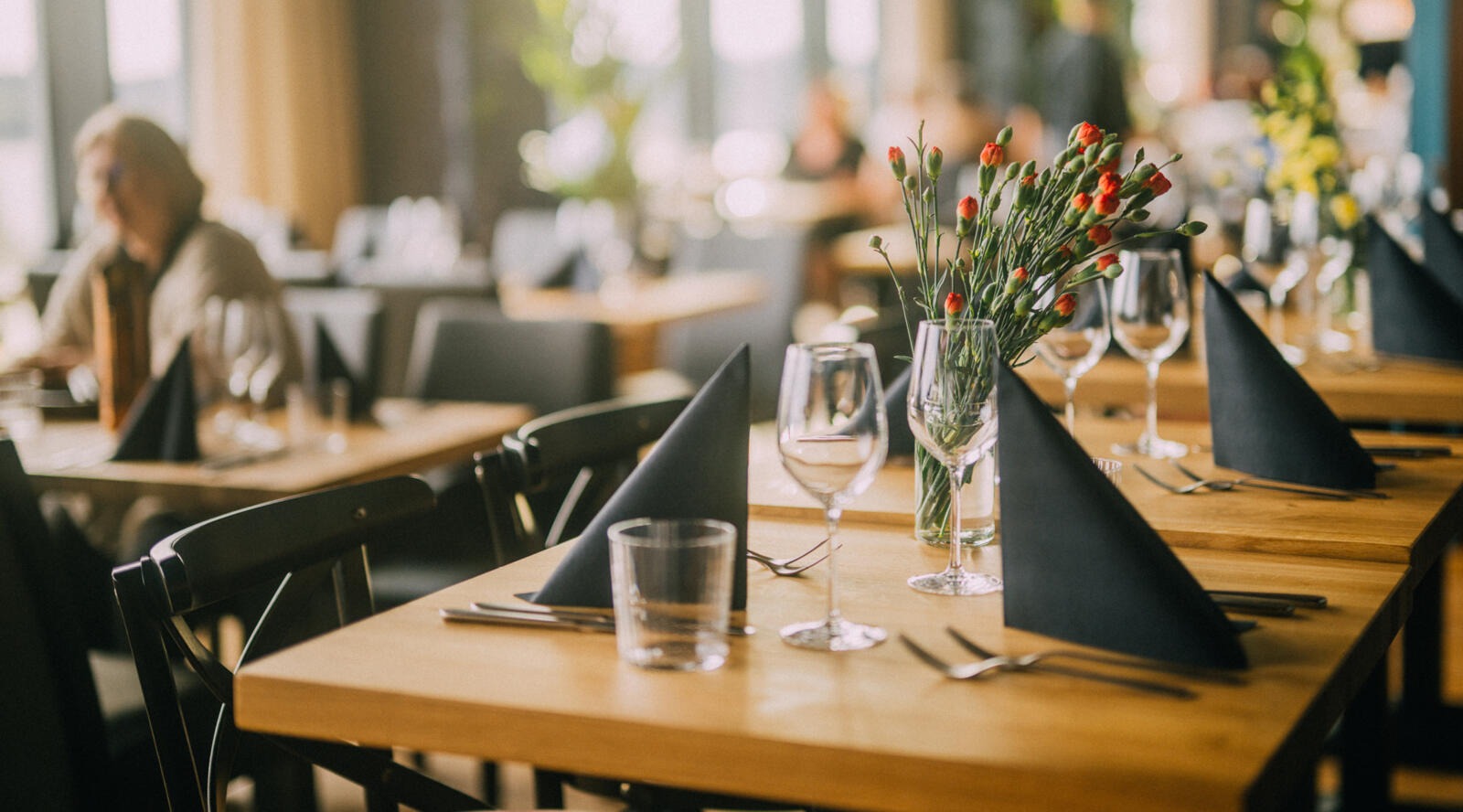 Elegant restaurangbord med röda blommor i en vas, vinglas och svarta servetter. Lokalen har träbord och svarta stolar.