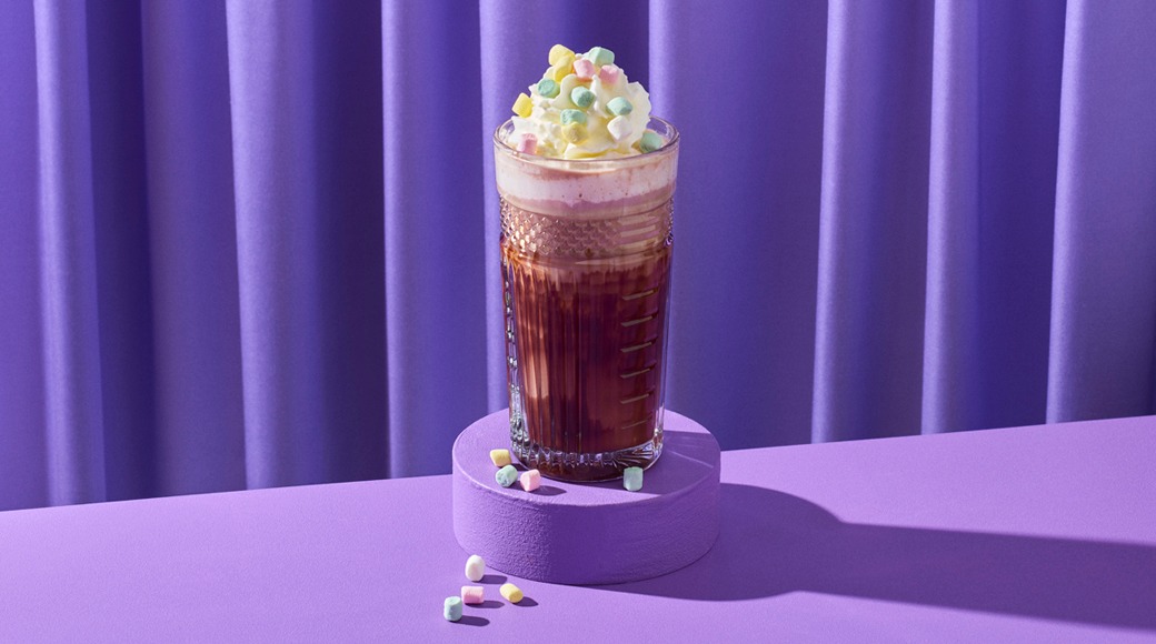 Kuumaa suklaata lasissa, päällä kermavaahtoa ja pastellinvärisiä minimarshmallowja. Lasi on violetilla korokkeella violettia verhoa vasten.