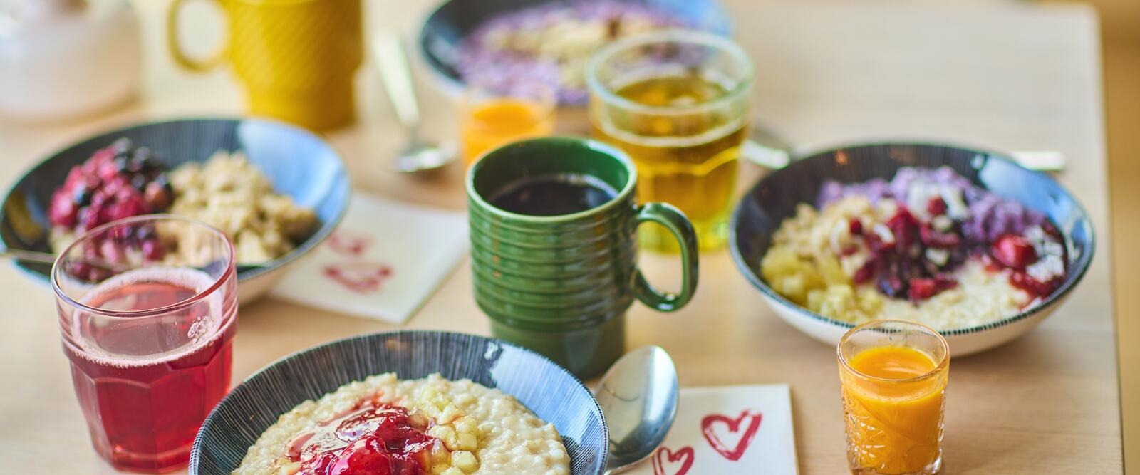 Breakfast table with porridge topped with berries, coffee, juice, and beer on a light wooden table.