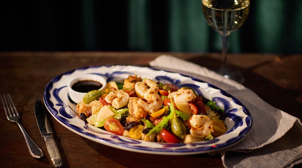 Värvilised krevetid ja köögiviljad sinise äärega taldrikul, valge veini klaas ja kastmekann laual nähtavad.