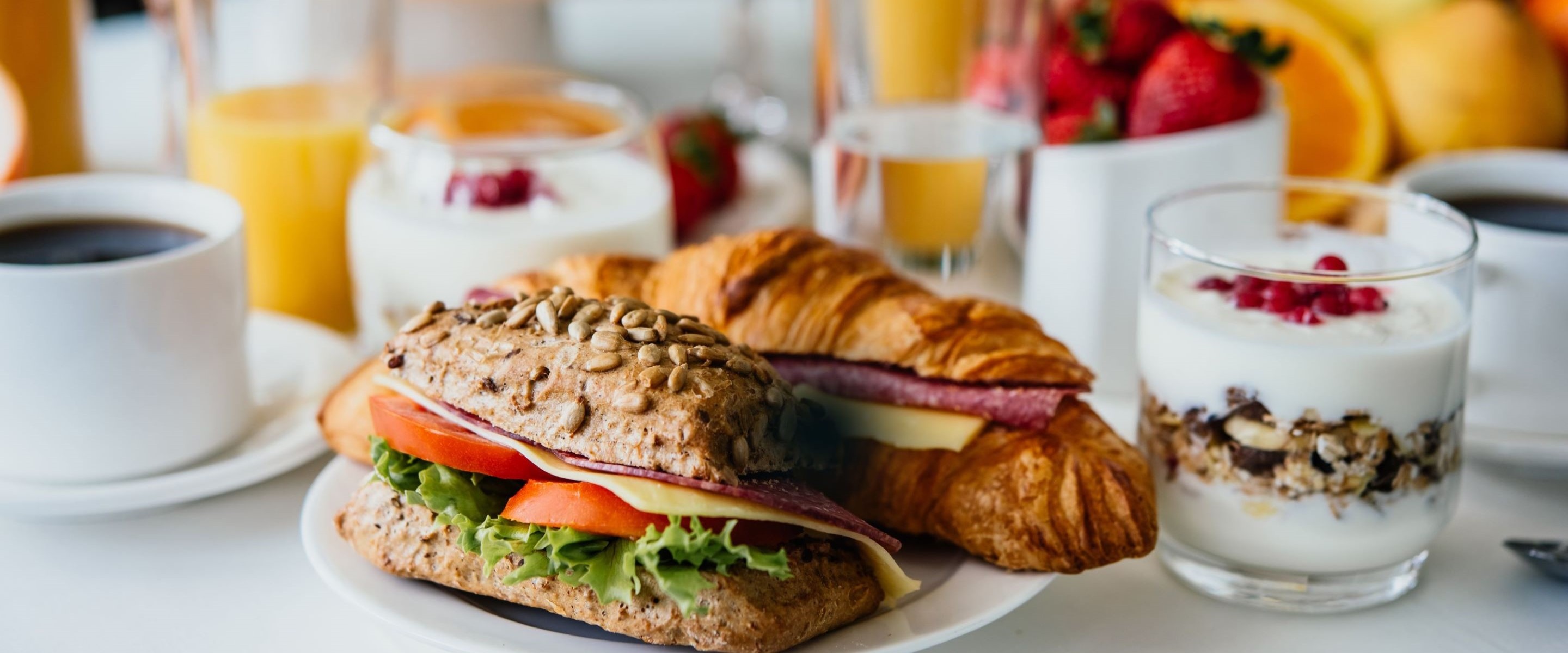 Runsas aamiaispöytä, jossa sämpylä, croissant, jogurttia, marjoja, mehua ja kahvia valkoisella pöydällä.