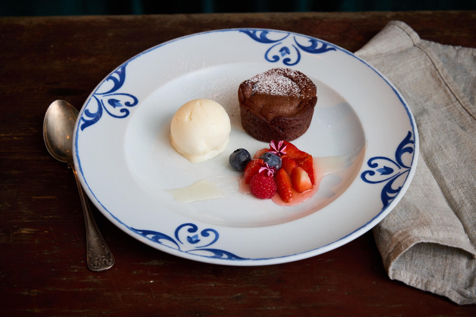 Valge taldrik siniste kaunistustega, millel šokolaadikook, vanillijäätisepall ja värsked marjad. Kõrval lusikas ja salvrätik.