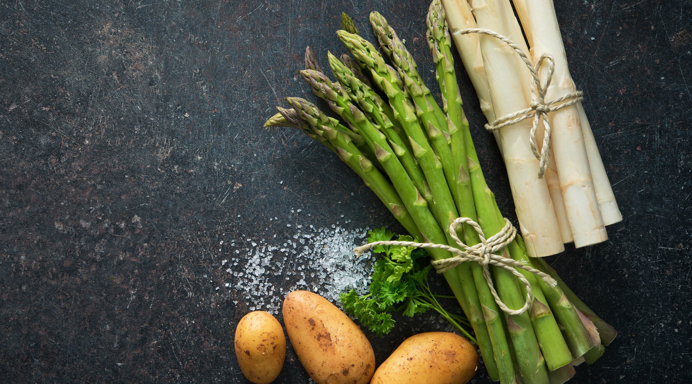 Vihreää ja valkoista parsaa narulla sidottuna, tuoreita perunoita ja persiljaa tummalla kivipinnalla ripoteltuna merisuolalla.