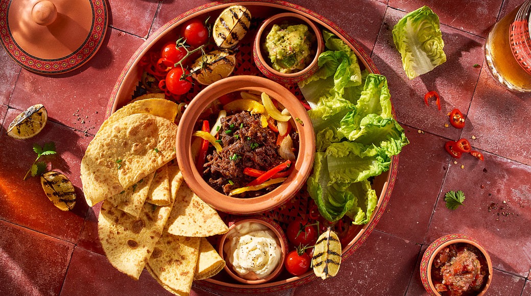 Terrakottakulhossa suikaloitua lihaa ja paprikaa, ympärillä tortillakolmioita, salaattia, guacamolea, smetanaa, tomaatteja ja grillattuja limetinpuolikkaita punaisella laatalla.