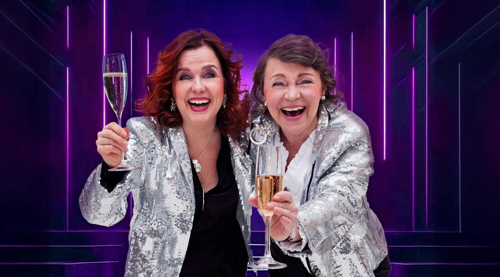 Två leende kvinnor i glittrande silverjackor skålar med champagneglas mot lila belysning.