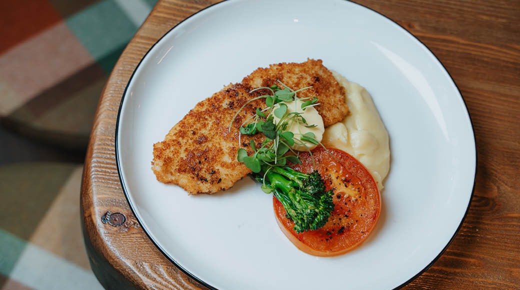 Vit tallrik på träbord med panerad kött eller fisk, potatismos, tomatskiva och grön broccoli garnerad med mikroörter.