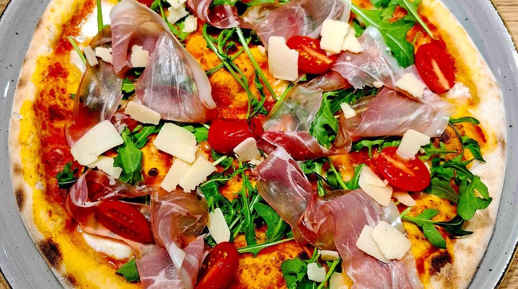 Gourmet-pizza täytetty prosciutto-kinkulla, parmesaanilastuilla, rucola-lehdillä ja kirsikkatomaateilla harmaalla lautasella.