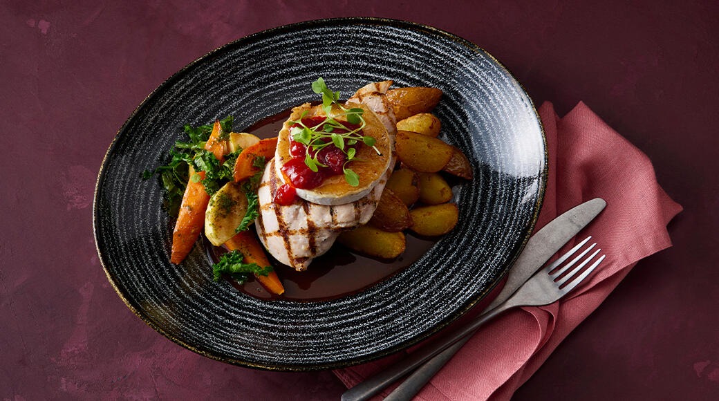 Grilled chicken with lingonberry jam, roasted carrots and potatoes on a dark plate. A red napkin and cutlery lie beside it.