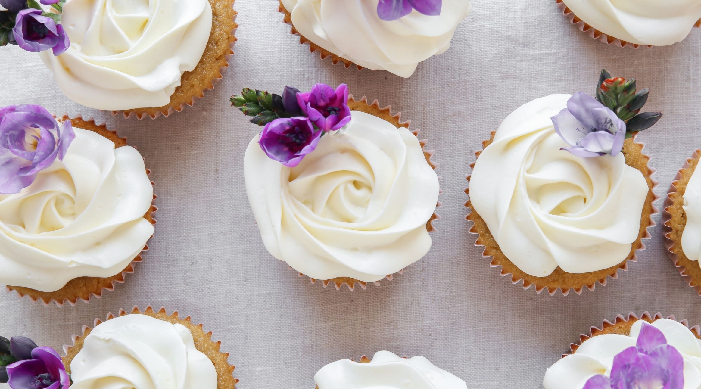 Kauniita kuppikakkuja, joissa on valkoista ruusun muotoista kuorrutetta ja lilan värisiä kukkakoristeita vaalealla kankaalla.