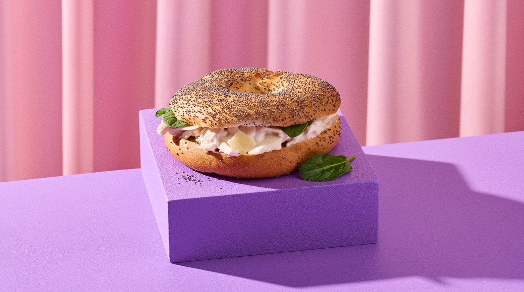 Unikkopäällysteinen bagel, jossa on kermaista täytettä ja pinaatinlehtiä, violetilla korokkeella vaaleanpunaista verhoa vasten.