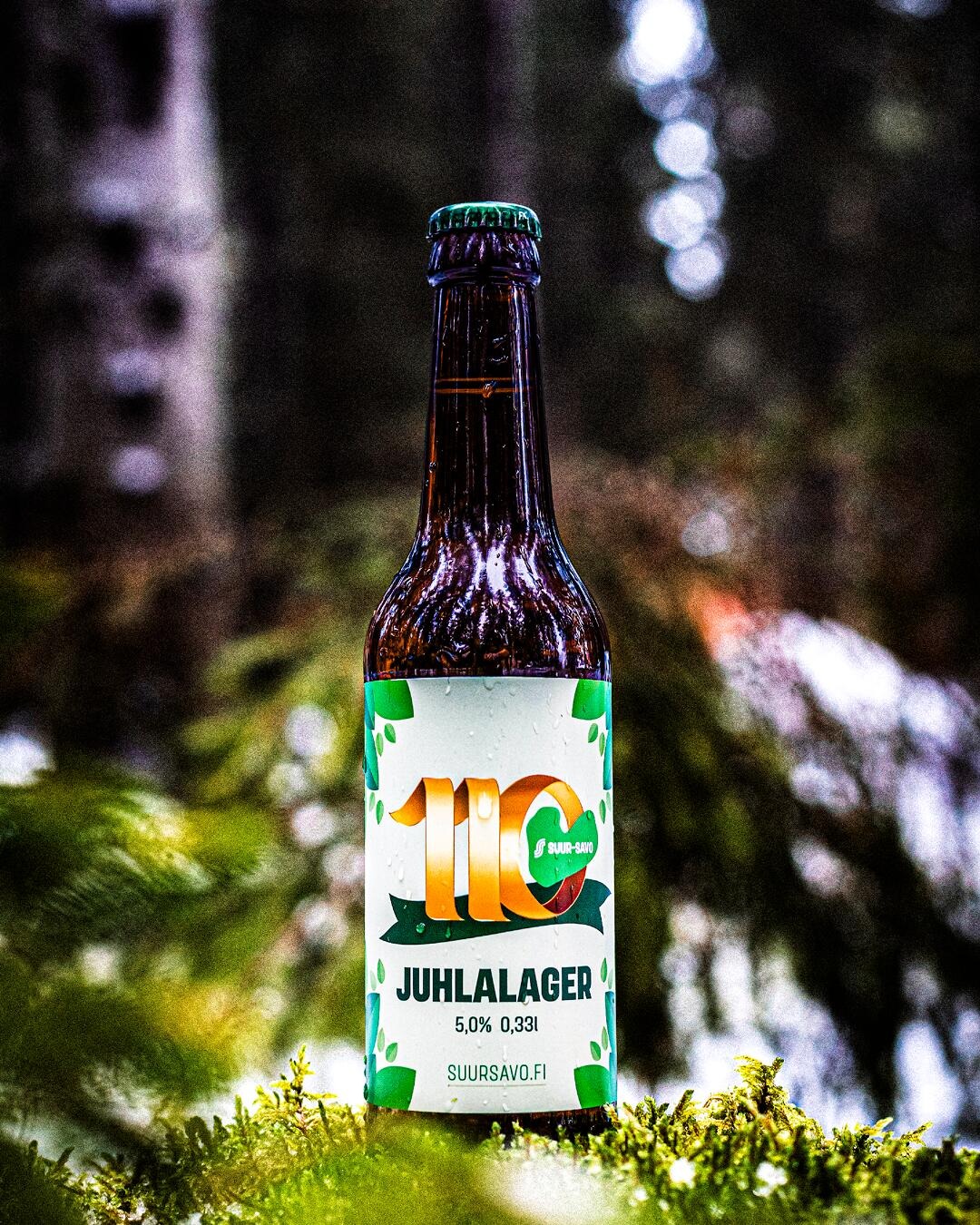 En brun glasflaska med Suur-Savo Juhlalagern på en mossig yta med dunkel natur och ljusglimtar i bakgrunden.