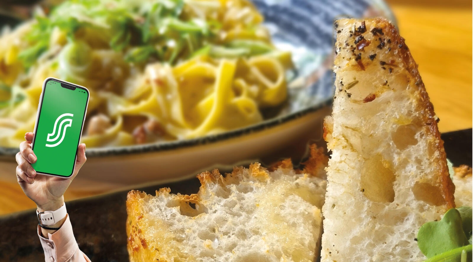 Hand holding a phone next to toasted bread and a pasta meal.