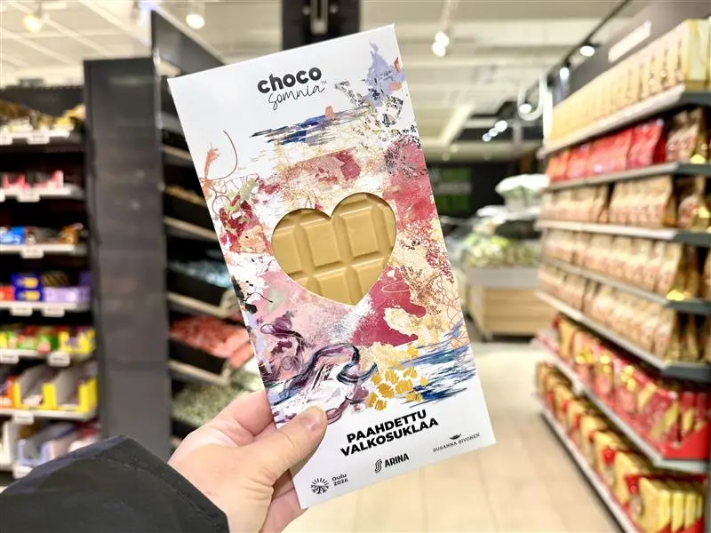Taustalla myymälä ja etualalla suklaalevy, jonka kuoressa Mielestäni-kuosi.