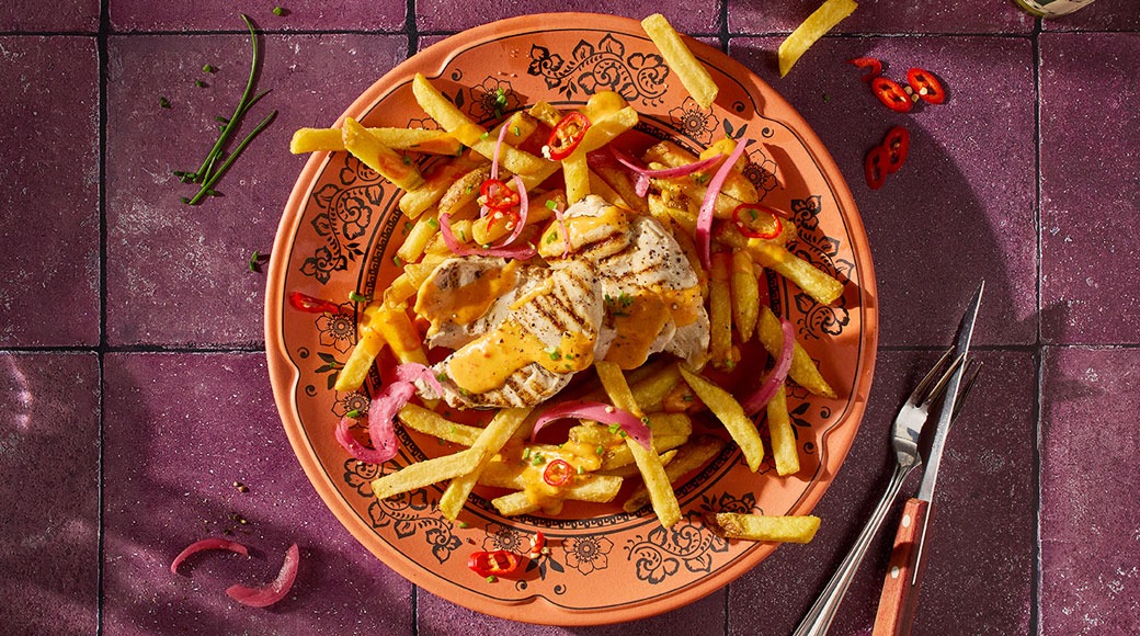 Terrakottalautasella grillattua kanaa, ranskalaisia perunoita, oranssia kastiketta, punasipulia ja chiliä. Vieressä haarukka ja veitsi violetilla laatoitetulla pöydällä.