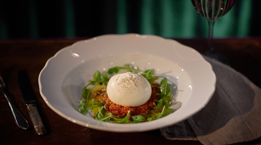 Valge portselantaldrik burrata juustupalli, tomatikastme ja värskete ürtidega. Taustal veinipokaal.