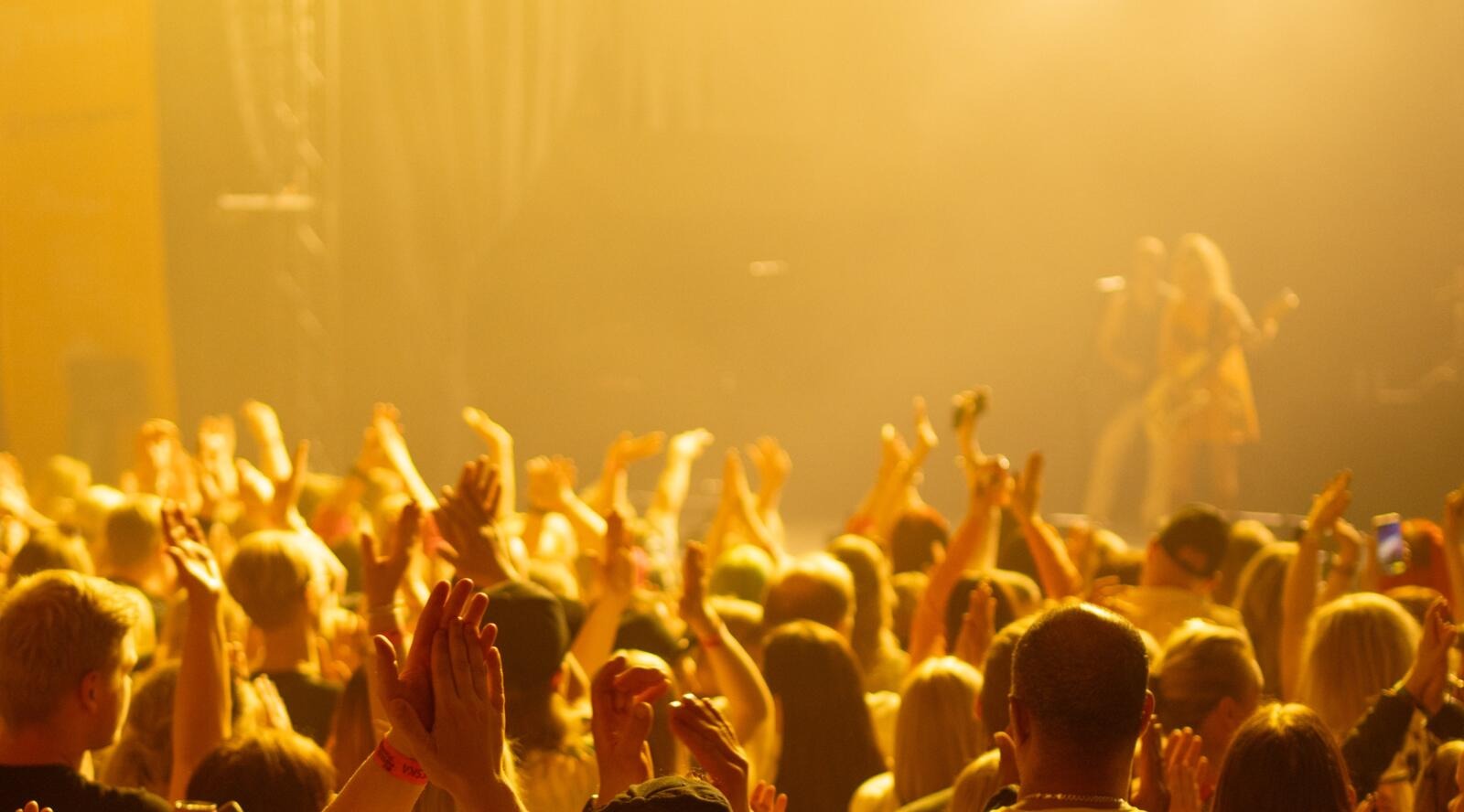 Concert crowd raising hands in the air under yellow lighting, with performer visible hazily on stage in the background.