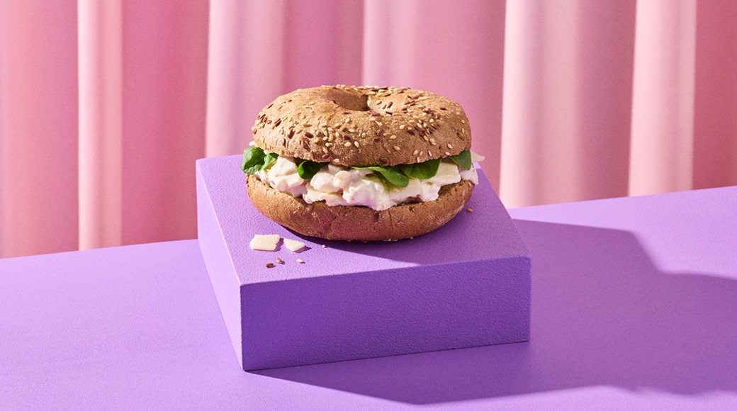 Siemenillä koristeltu täysjyväbagel, jonka välissä on tuorejuustoa, pinaatinlehtiä ja juustoa. Bagel on violetin korokkeen päällä vaaleanpunaisen verhon edessä.