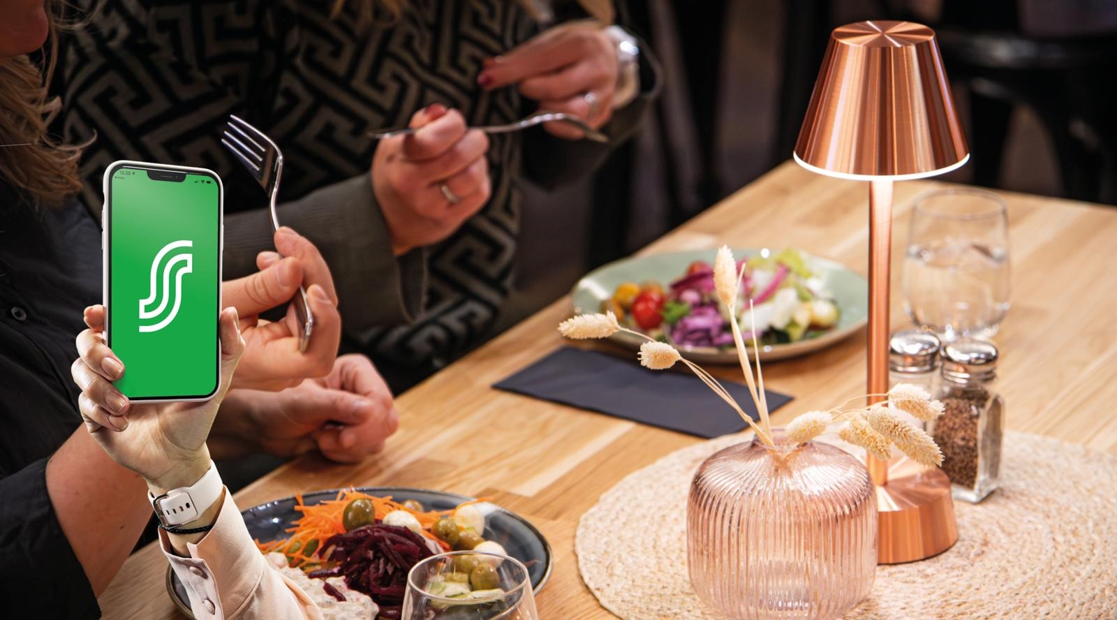 Person showing smartphone at restaurant table. Table has colorful salads, copper table lamp and decorative items.