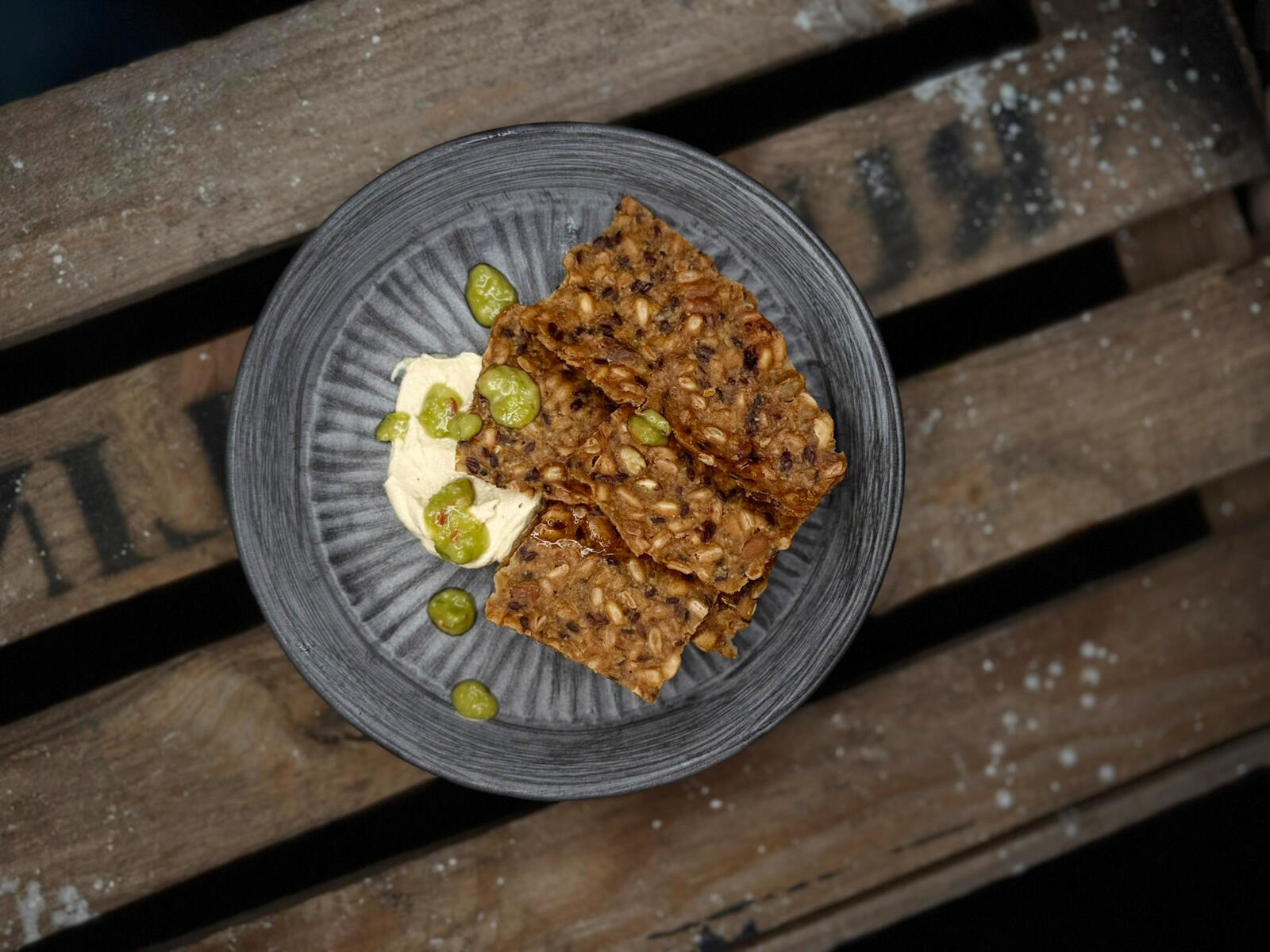 Seeded crackers, hummus dip, and green olives on a gray plate resting on wooden planks.