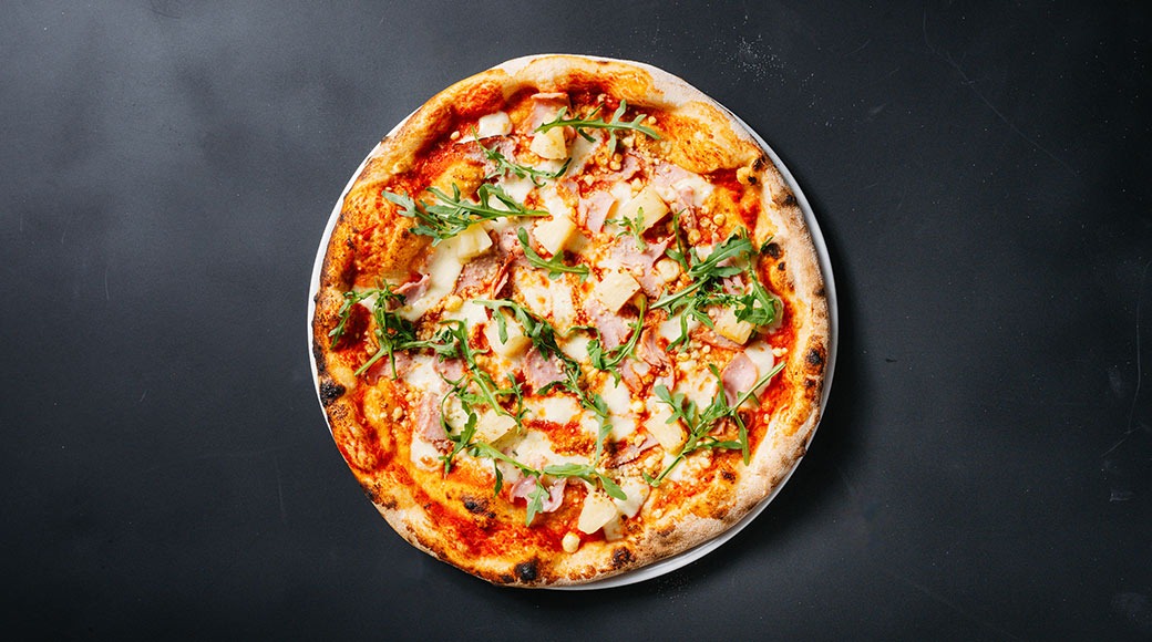 Pizza kinkkua, juustoa ja rucola-lehtiä tummanharmaalla alustalla.