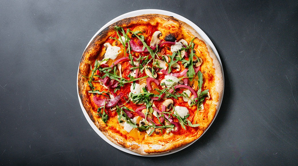 Vegepizza, jossa on herkkusieniä, rucola-salaattia ja juustoa tummalla alustalla.