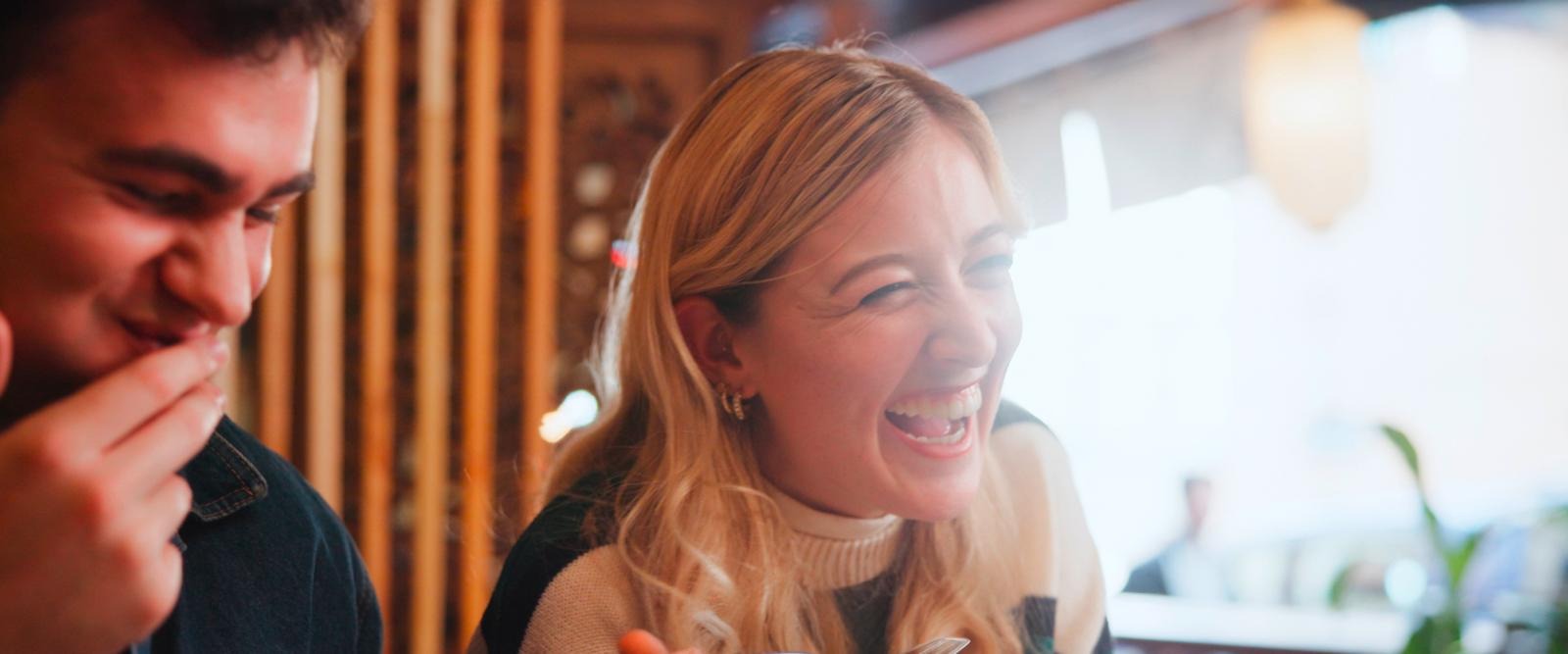 Kaksi ihmistä nauraa yhdessä lämpimästi valaistun ravintolan tai kahvilan pöydässä. Taustalla näkyy puinen sisustus.