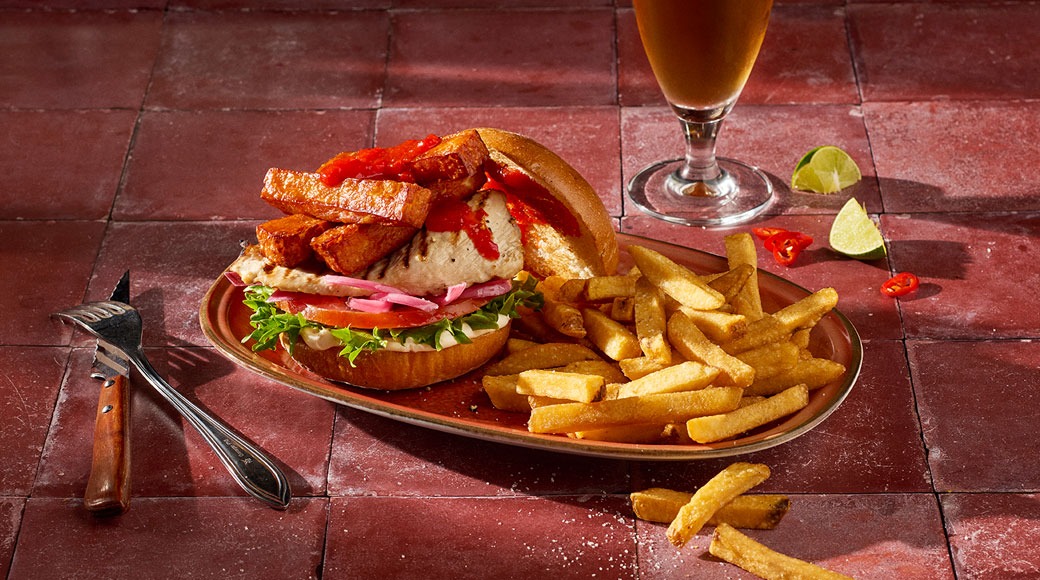 Grillitud kanaburger, mille peal on friteeritud ribad ja punane kaste. Taldrikul on friikartulid, kõrval õlleklaas, laimitükid ja kahvel punasel plaatlaual.