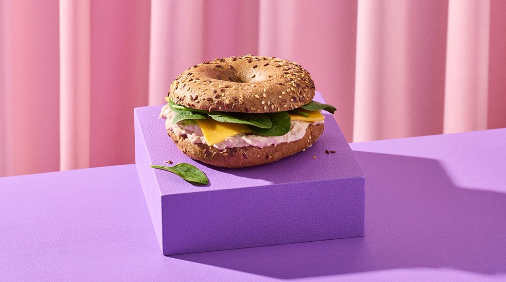 Siemenillä koristeltu bagel, jonka välissä on tuorejuustoa, juustoviipale ja pinaatinlehtiä. Bagel on violetin korokkeen päällä vaaleanpunaista verhoa vasten.