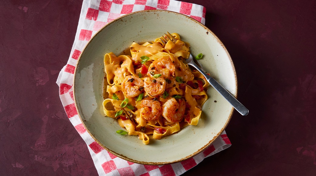 Bowl of tagliatelle with shrimp and creamy tomato sauce, topped with spring onion. The bowl sits on a red and white checkered cloth.