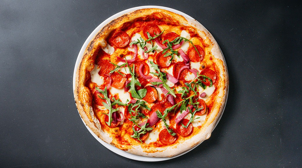 Pizza valkoisella lautasella tummalla alustalla, päällä salamia, kinkkua, juustoa ja rucolaa.