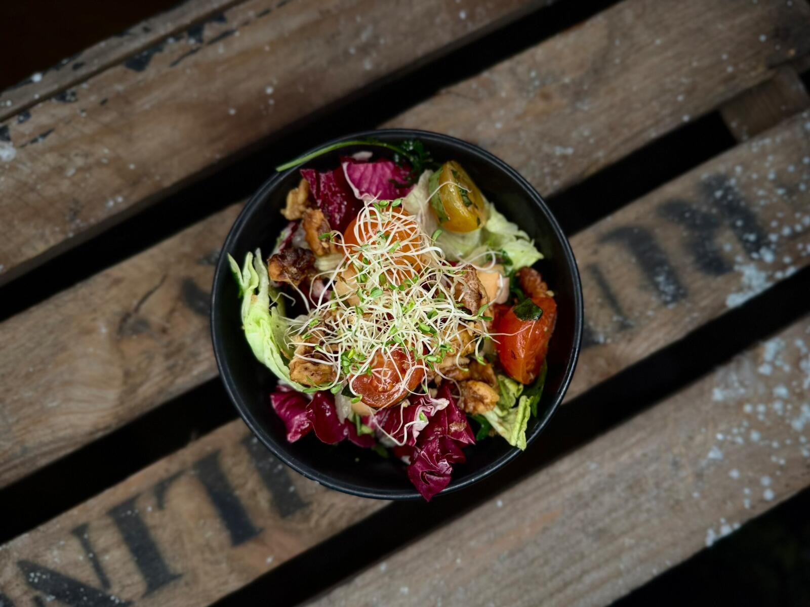 Colorful salad in a black bowl on wooden surface. Contains sprouts, red cabbage, tomato, and green lettuce.
