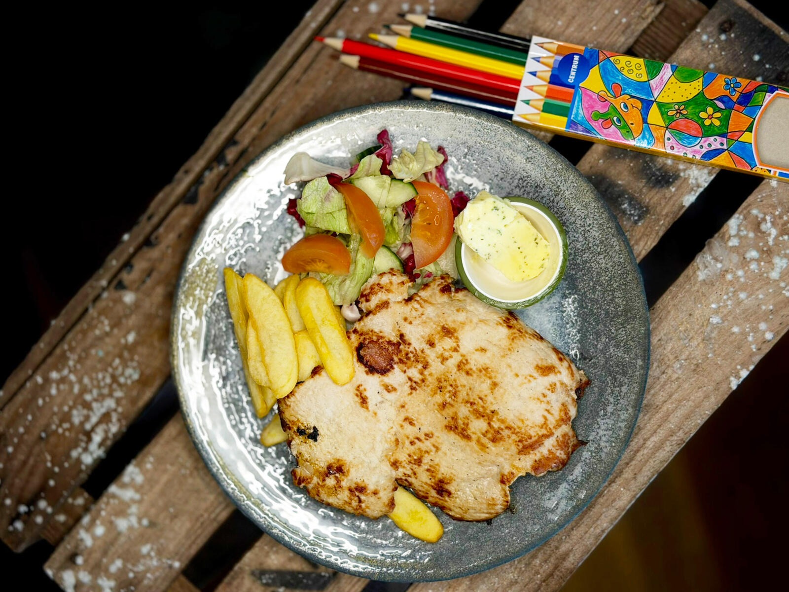 Gray plate on wooden surface with grilled meat, french fries, salad, tomatoes and a pat of butter. Colorful pencils rest nearby.
