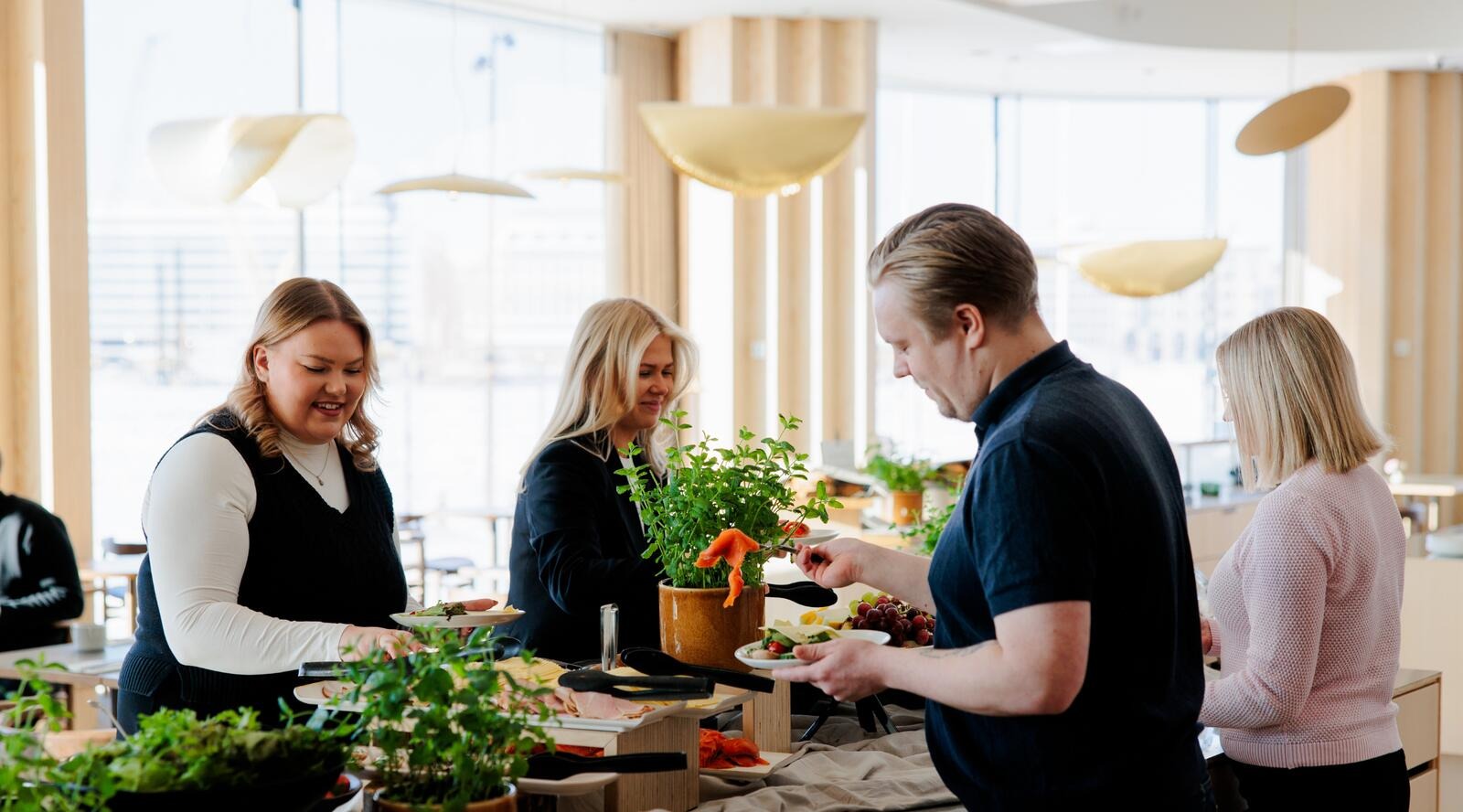 Ihmiset ottamassa ruokaa noutopöydästä valoisassa ravintolassa. Pöydällä raikkaita yrttejä ja erilaisia ruokavaihtoehtoja.