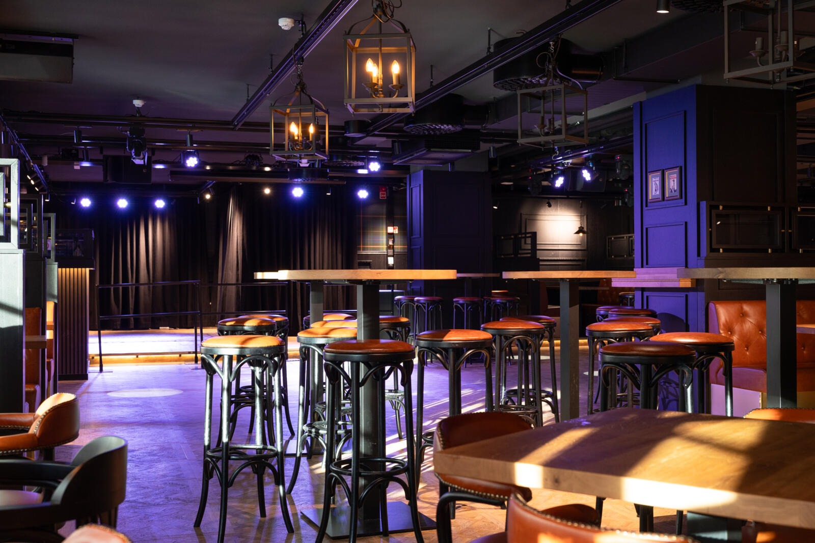 Baari tai keikkatila, jossa on korkeat pöydät ja baarituolit sekä pieni lava taustalla. Valaistu vintage-tyylisillä lyhtyvalaisimilla ja kohdevaloilla.