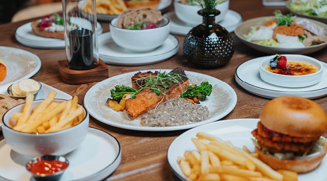 Wooden table filled with various food dishes, including a burger, french fries, and a main course with vegetables and sauce.