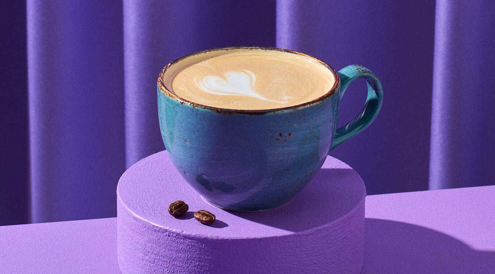 Blå keramikkopp med kaffe och ett hjärtmönster i skummet. Koppen står på en lila sockel med två kaffebönor bredvid.
