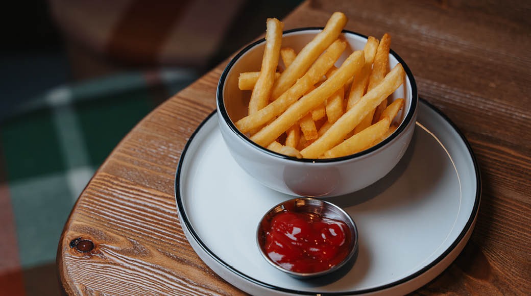 Rapeita ranskalaisia perunoita valkoisessa kulhossa ja ketsuppia pienessä astiassa valkoisella lautasella puisella pöydällä.