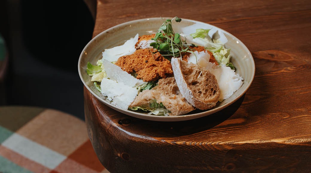 Salaattiannos valkoisessa kulhossa sisältäen leivänpaloja, rapean pinnoitettua proteiinia ja tuoreita vihanneksia puisella pöydällä.