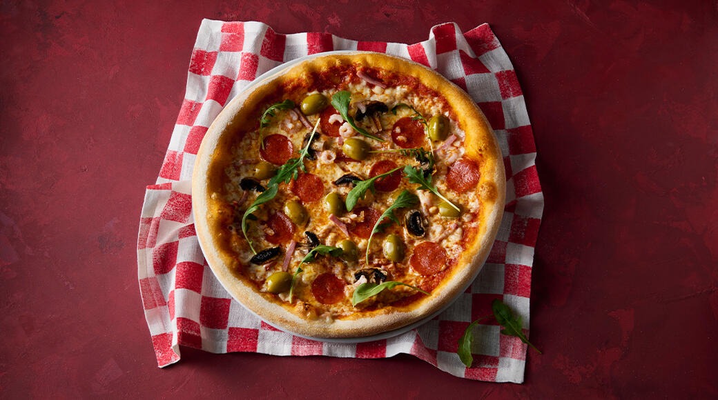 Pizza topped with pepperoni, green olives, mushrooms and rocket. It sits on a red and white checkered cloth on a red surface.