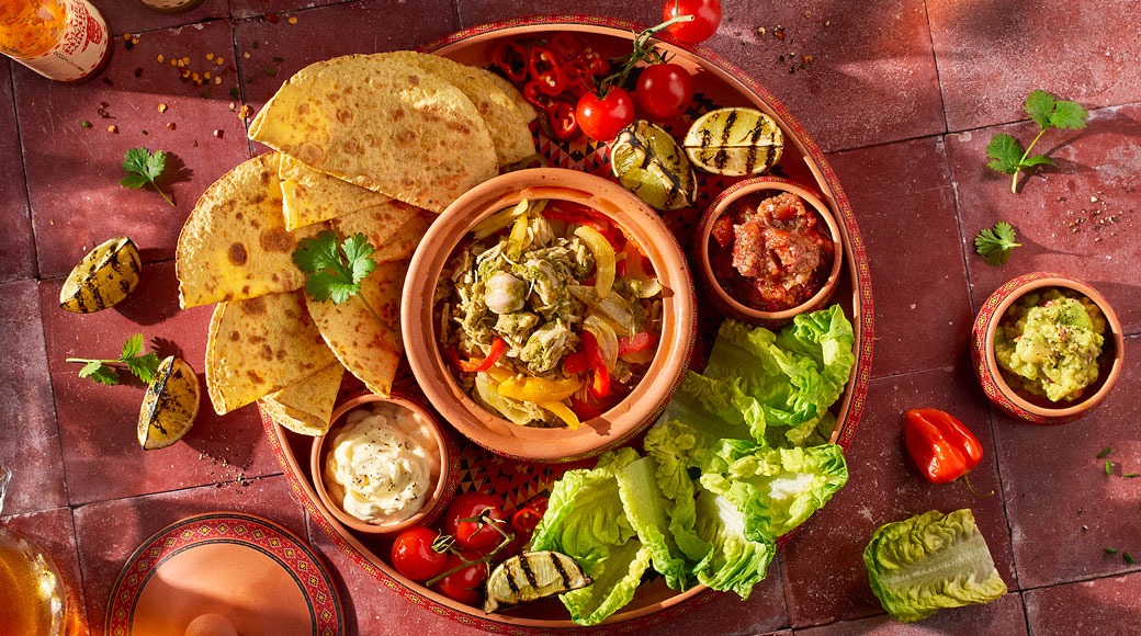 Yläkuva tarjottimesta, jolla on tortillakolmioita, kulhollinen paistettua kanaa ja paprikaa, salsaa, guacamolea, smetanaa, salaattia, kirsikkatomaatteja ja grillattuja lime-puolikkaita.