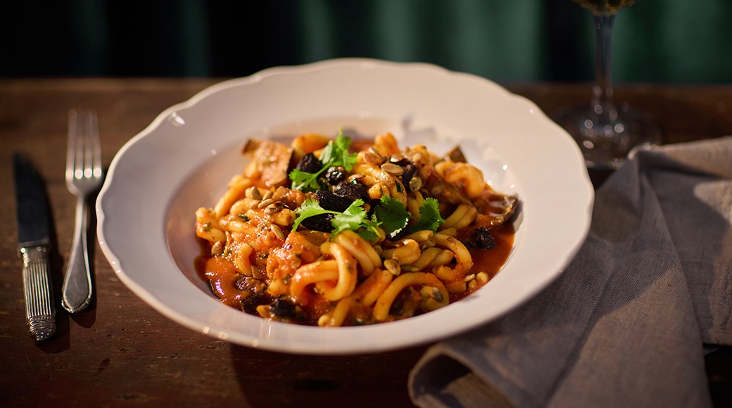 En vit tallrik med pasta i tomatsås, garnerad med färska örter och nötter på ett träbord.