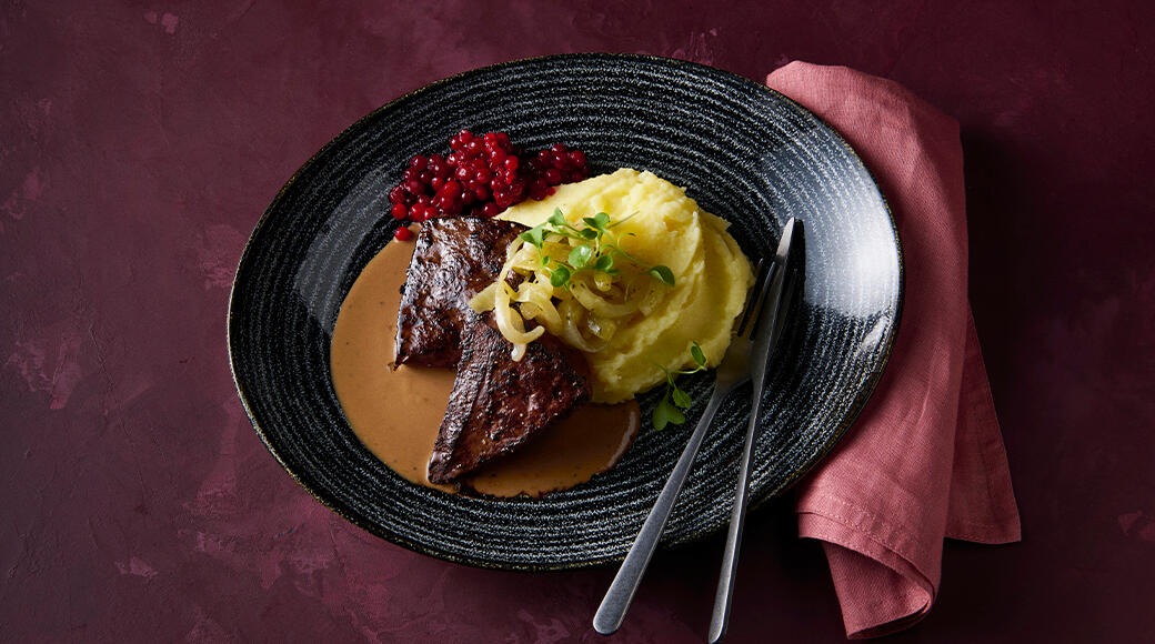 Liver steaks in gravy with mashed potatoes, caramelised onions and lingonberries on a black plate. A fork, knife and red napkin lie beside it.