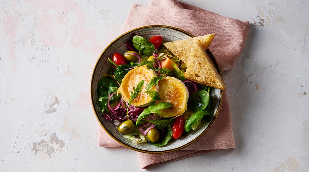Salad bowl seen from above, with leafy greens, tomatoes, olives, red onion and baked goat cheese, plus toasted bread triangles.