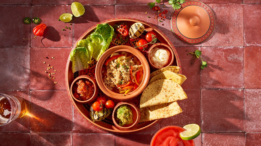 Terrakotataldrik tortillade, paprikaga hakitud liha, salsa, guakamole, hapukoore ja salatiga. Kõrval laimiviilud ja joogid.