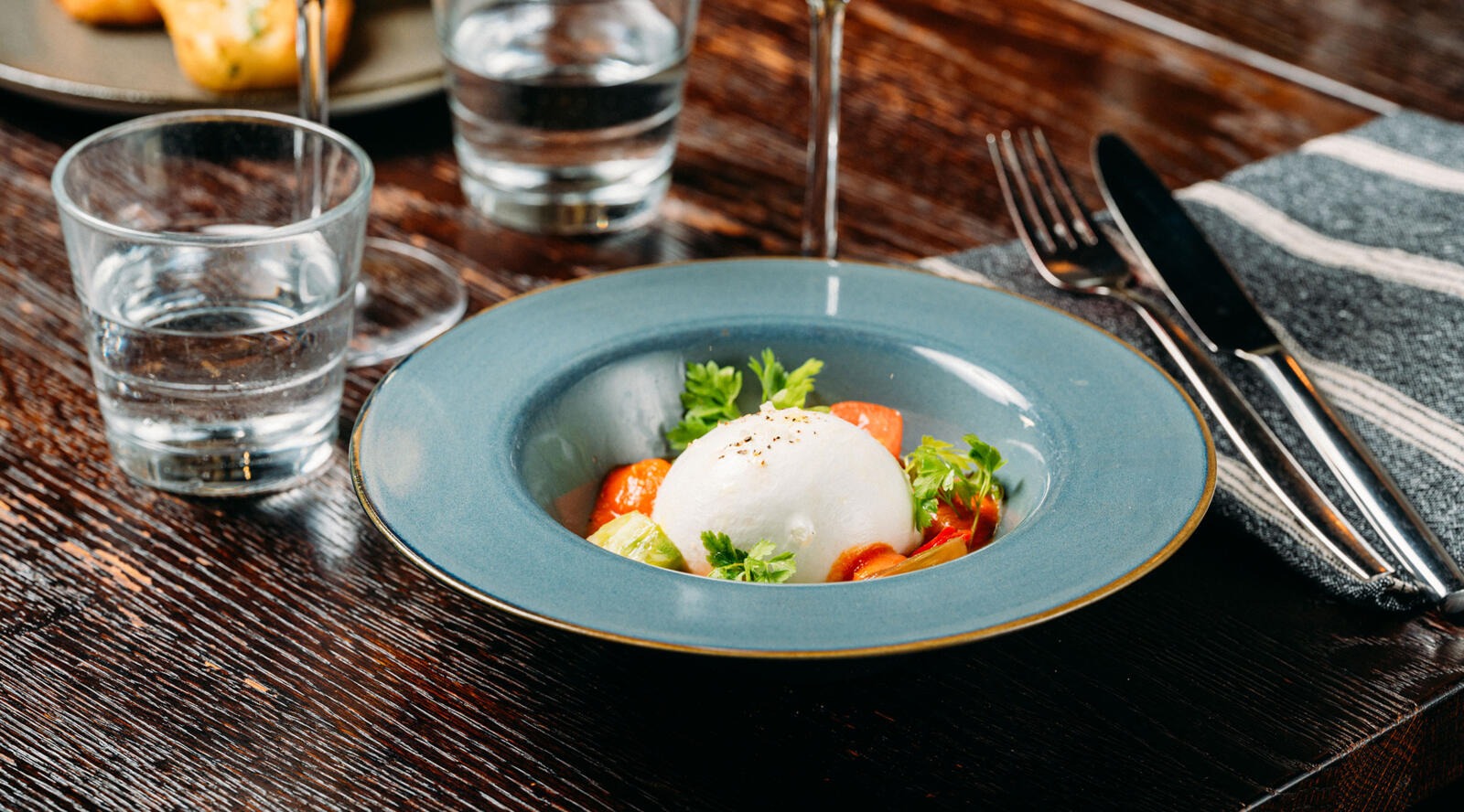 Sinisellä lautasella tarjoiltu burrata-juusto kirsikkatomaattien ja tuoreiden yrttien kera puisella pöydällä.
