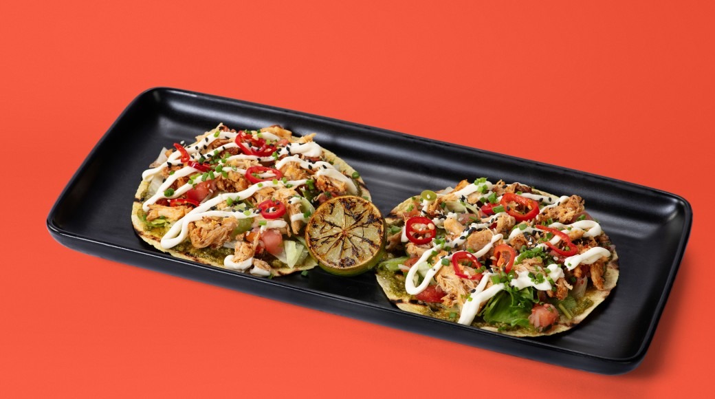 Three tacos on a black plate, filled with grilled chicken, sautéed peppers, green lettuce and white sauce, with a grilled lime wedge on the side.