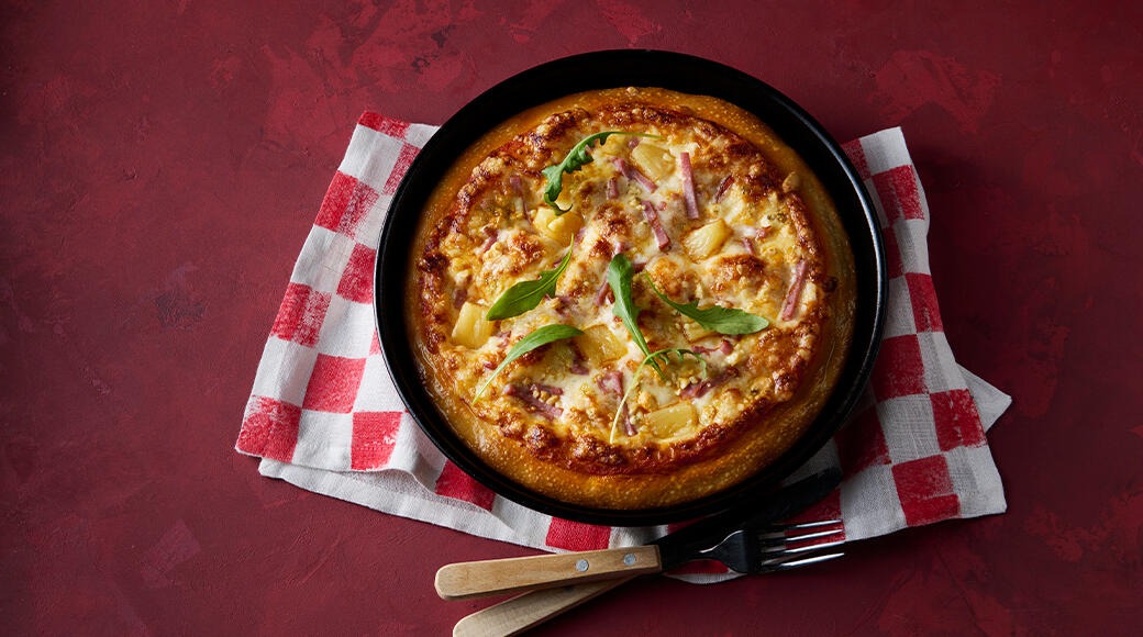 Uunituore ananas-kinkkupizza mustalla pellillä, päällä ruukolaa. Pelti on punavalkoruutuisen liinan päällä, vieressä haarukka ja veitsi.