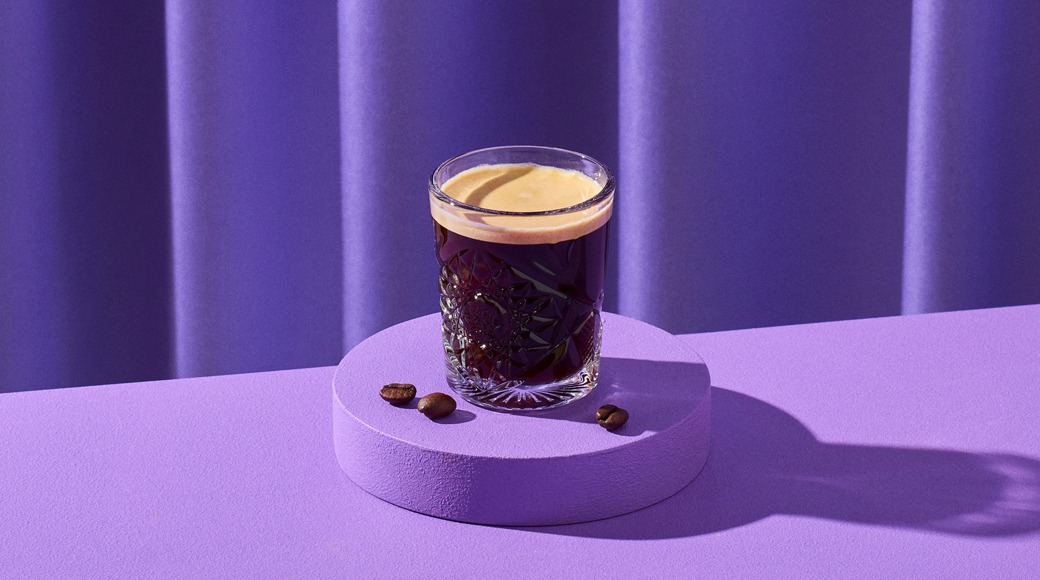 Lasillinen espressoa kristallikuvioidussa lasissa violetilla korokkeella. Vieressä kahvipapuja, taustalla violetti verho.