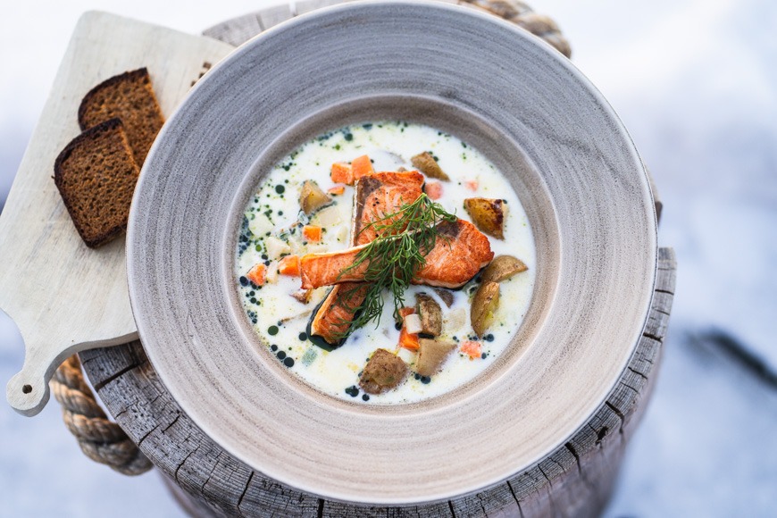 Lautasellinen kalakeittoa, jossa on lohta, tilliä ja vihanneksia. Vieressä tummaa leipää puulaudalla.