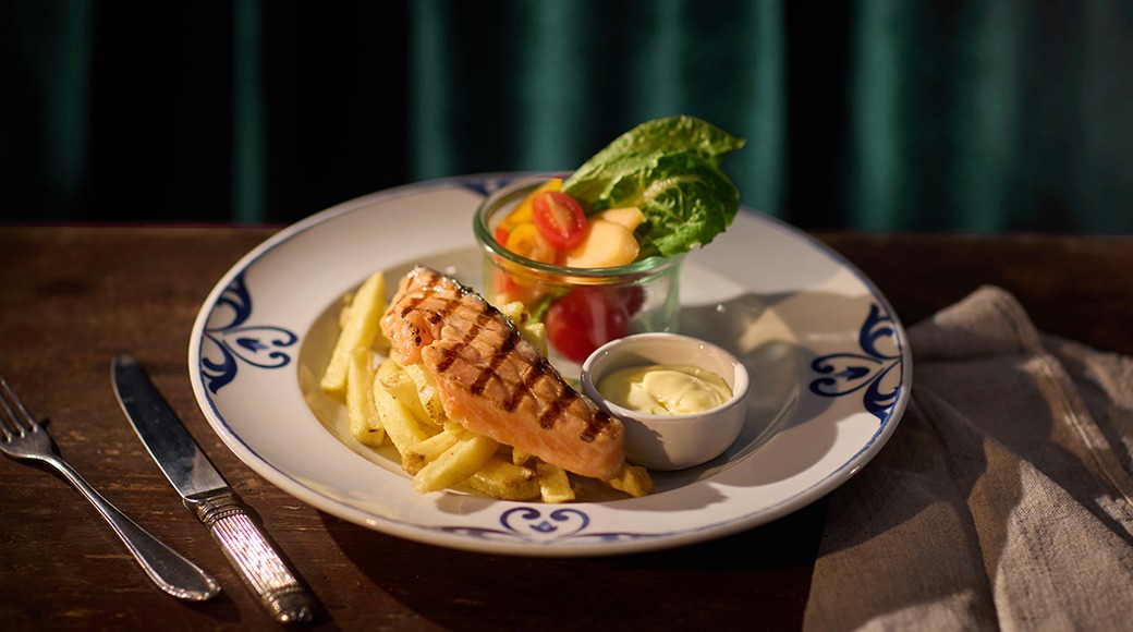 Annos grillattua lohta, ranskalaisia perunoita, majoneesia pienessä kupissa ja salaattia lasissa. Koristeellinen sinivalkoinen lautanen puisella pöydällä.